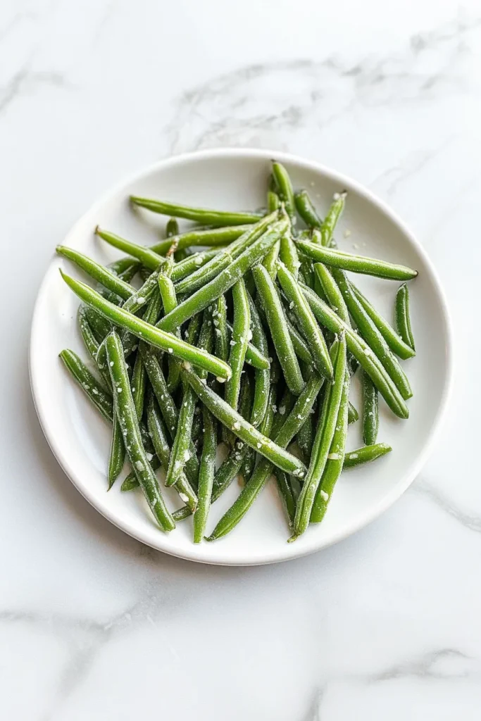 Glossy green beans piled on a white plate, sprinkled with coarse salt.